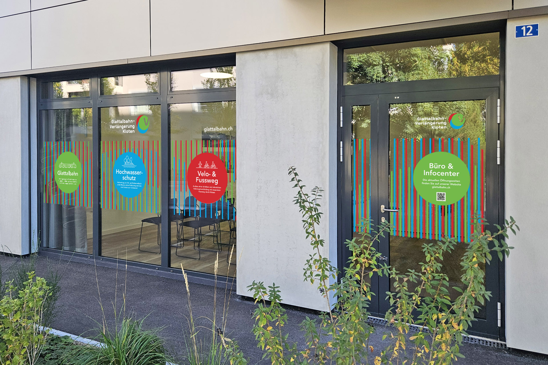 Eingang mit Glasfront des Büro & Infocenters mit bunten Aufklebern und Hausnummer 12, im Vordergrund grüne Bepflanzung.