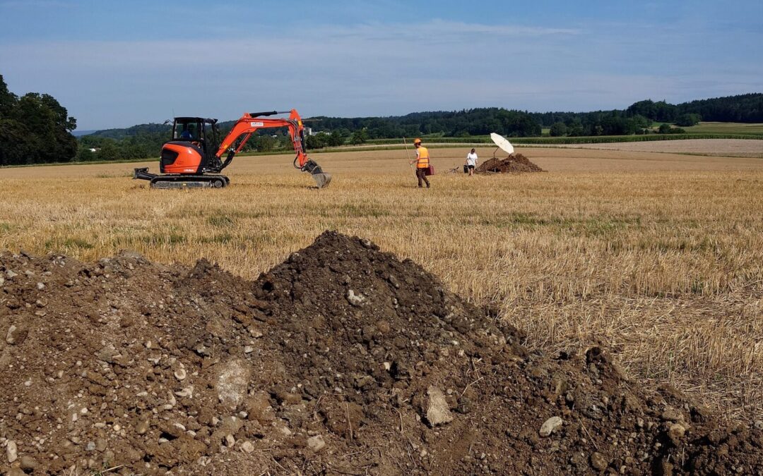 Ein Schaufelbagger steht auf einem gemähten Weizenfeld in Kloten, beobachtet von einem Archäologen und bereit, einen Teil des Bodens auszuheben.