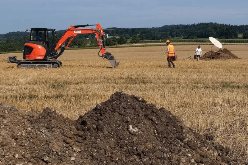 Ein Schaufelbagger steht auf einem gemähten Weizenfeld in Kloten, beobachtet von einem Archäologen und bereit, einen Teil des Bodens auszuheben.