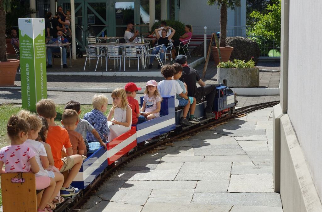 Kinder sitzen auf einer Miniatureisenbahn mit blau-weiss-rotem Muster, die auf einem gepflasterten Weg fährt