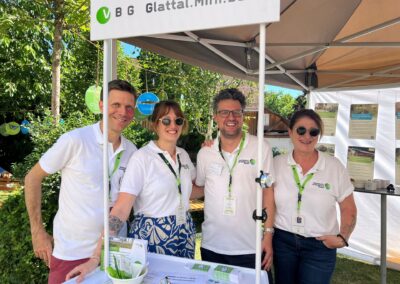 Vier Personen in weissen Poloshirts stehen lächelnd an einem mit «VBG Glattal.Mini.Bahn» beschrifteten Stand.