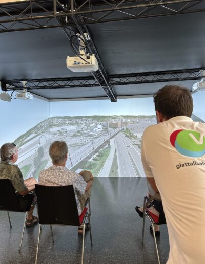 3 Personen sitzen und schauen auf ein 3D-Modell, 1 Mann im weissen Poloshirt mit Logo präsentiert. 3 Projektoren an der Decke