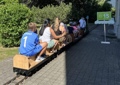 Mehrere Personen sitzen auf einer Miniatureisenbahn, ein Schild mit «15» symbolisiert den Viertelstundentakt der Glattalbahn.